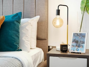 Un lit bien fait, avec des oreillers turquoise et blancs, une table de chevet sur laquelle se trouve une lampe, une petite plante et un appareil numérique affichant un menu.