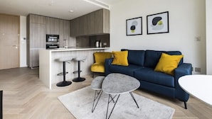 A modern living room with a blue sofa, two yellow cushions, a grey coffee table, and a kitchen in the background.