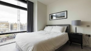 A modern hotel room with a large bed, a bedside table with a lamp, a framed picture on the wall, and a view of the city through a large window.