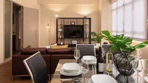 A modern living room with a sofa, dining table set for four, a TV mounted on a wall, and a large window. - Lovely apartment for 2 people with WIFI and TV (London)