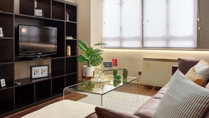 A modern living room with a flat-screen TV, dark wooden shelving, a glass coffee table, a large window, and a radiator. - Lovely apartment for 2 people with WIFI and TV (London)
