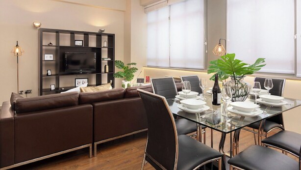 A modern living room with a brown sofa, a glass dining table set for four, and a flat-screen TV mounted on the wall. - Lovely apartment for 2 people with WIFI and TV (London)