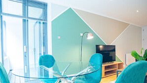 Un soggiorno moderno con un tavolo da pranzo in vetro, sedie color petrolio, una TV a schermo piatto e una parete verde a contrasto.