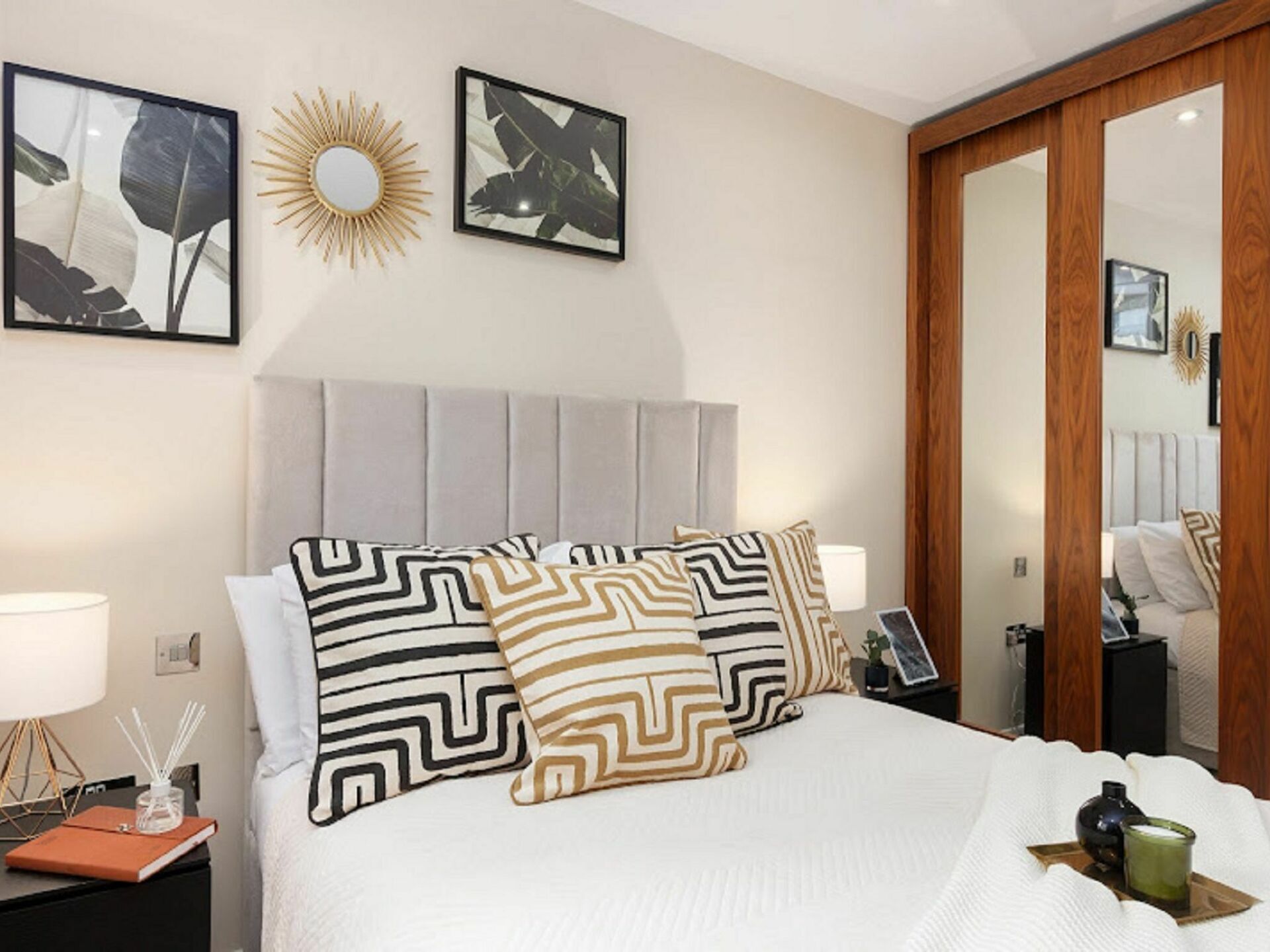 Habitación de hotel impecable con una cama grande, una mesita de noche con una lámpara, otro mueble con un libro y una vela, y un espejo en la pared.