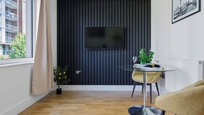A modern room with a flat-screen TV mounted on a dark wall, a dining table with chairs, a potted plant, and a window with curtains. - Lovely apartment for 2 guests with WIFI, TV and balcony (London)