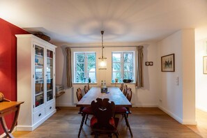 A dining room with a wooden table, red upholstered chairs, a white cabinet with glass doors, and two windows with curtains.