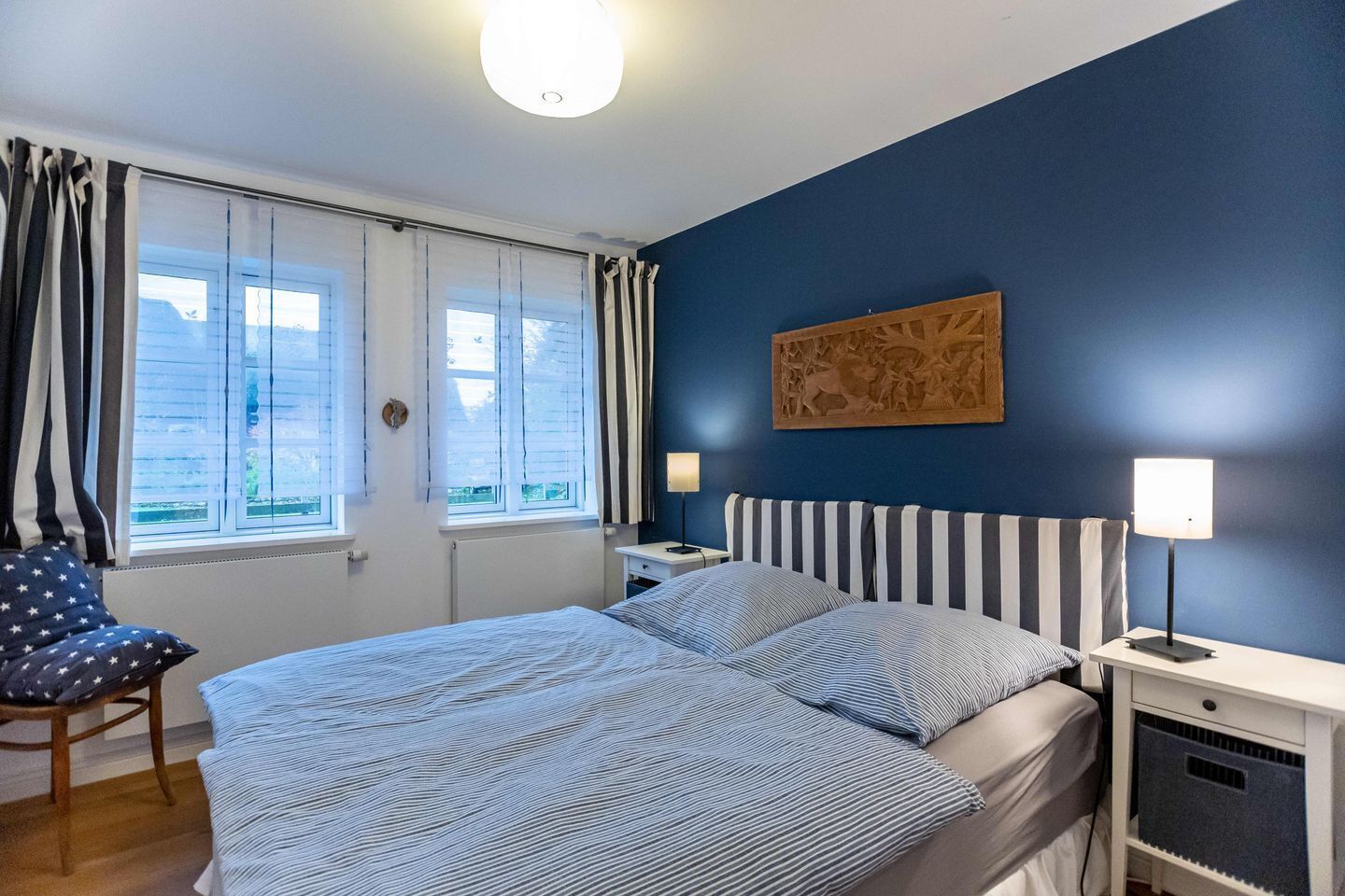 A bedroom with a bed, a chair, a nightstand, a lamp, and a window with curtains.