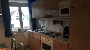 Une cuisine compacte équipée d’une cuisinière, d’un évier et d’un four à micro-ondes. On y trouve une fenêtre avec des rideaux, un radiateur et une petite plante.