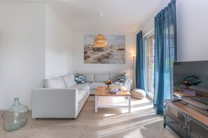 Ein modernes Wohnzimmer mit einem hellgrauen Sofa, einem Couchtisch aus Holz mit Büchern und einer Obstschale, einer hängenden Lampenschirm aus Bändern und einem großen Fenster mit blauen Vorhängen.