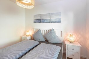 Ein Schlafzimmer mit zwei Einzelbetten, einem weißen Kopfstück, einem Nachttisch mit Lampe und einem an der Wand hängenden Gemälde.