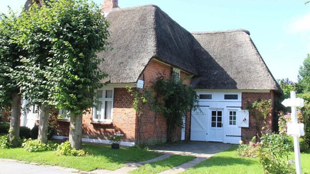 Een klassiek vakantiehuis met rieten dak, een witte voordeur en een kleine tuin met een boom en een bord.