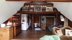 Een loft-achtige kamer met een houten plafond, een zithoek met een houten tafel en stoelen, een bank met gestreepte kussens en een trap naar een verdieping erboven.