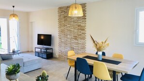 A modern living room with a flat-screen TV, a sofa, a dining table with chairs, and a stone accent wall. - LocaJard House "Les Sternes" 400m from sandy beaches, 70m², 3 bedrooms (Saint-Vincent-sur-Jard)
