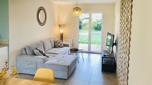 A modern living room with a grey sofa, a yellow chair, a TV on a stand, and a large window with a view of greenery. - LocaJard House "Les Sternes" 400m from sandy beaches, 70m², 3 bedrooms (Saint-Vincent-sur-Jard)