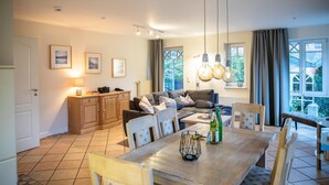 Ein gut beleuchteter Wohnbereich mit Sofa, Esstisch und Stühlen. An der Wand hängen Bilder in Rahmen, ein Schrank und ein Fenster mit Vorhängen.