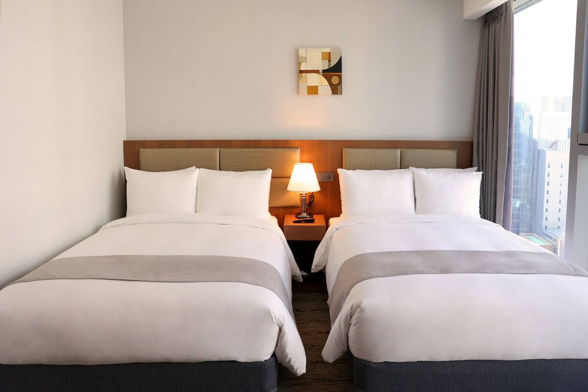Ein Hotelzimmer mit zwei Betten, einem Nachttisch mit Lampe, einem Wandbild und Blick auf die Stadt.