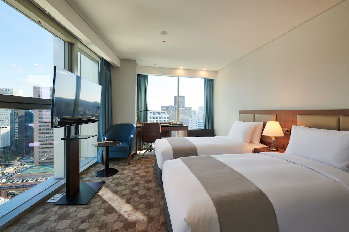 Ein Hotelzimmer mit zwei Betten, einem Schreibtisch, einem Stuhl, einem Fernseher und einem großen Fenster mit Blick auf die Stadt.