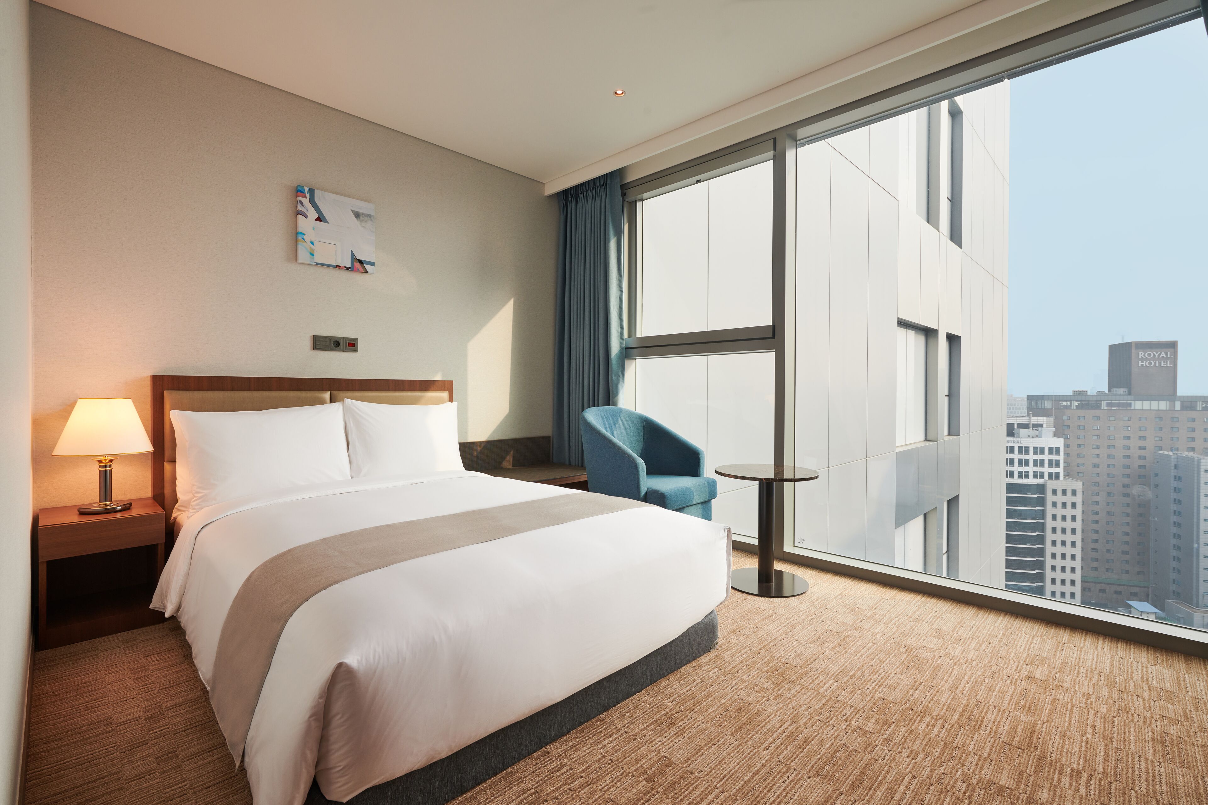A hotel room with a large bed, a desk with a lamp, a chair, and a view of the city through large windows.