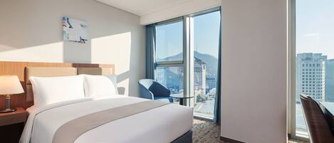 A modern hotel room with a large bed, a desk, a chair, and a view of the city through large windows.