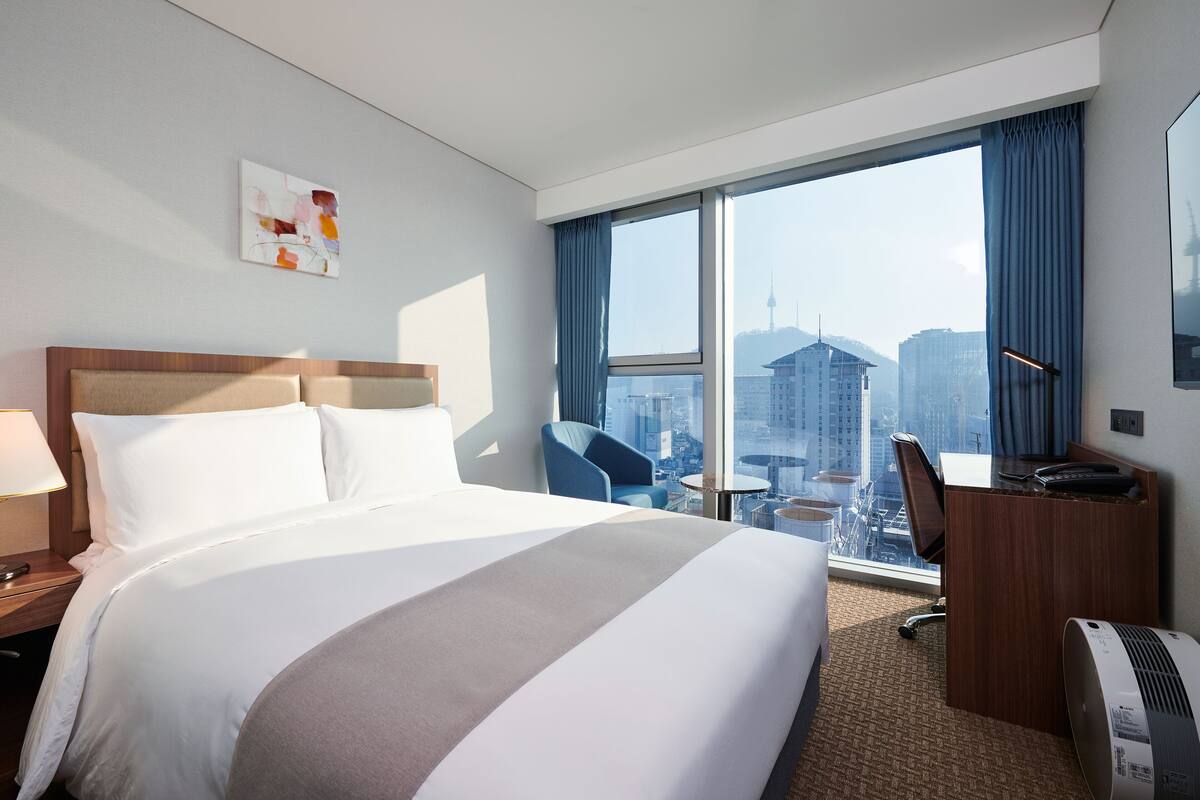 Ein Hotelzimmer mit einem großen Bett, einem Schreibtisch, einem Sessel und Blick auf die Stadt.