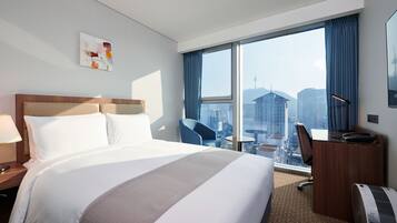 A hotel room with a large bed, a desk, a chair, a lamp, and a view of the cityscape.