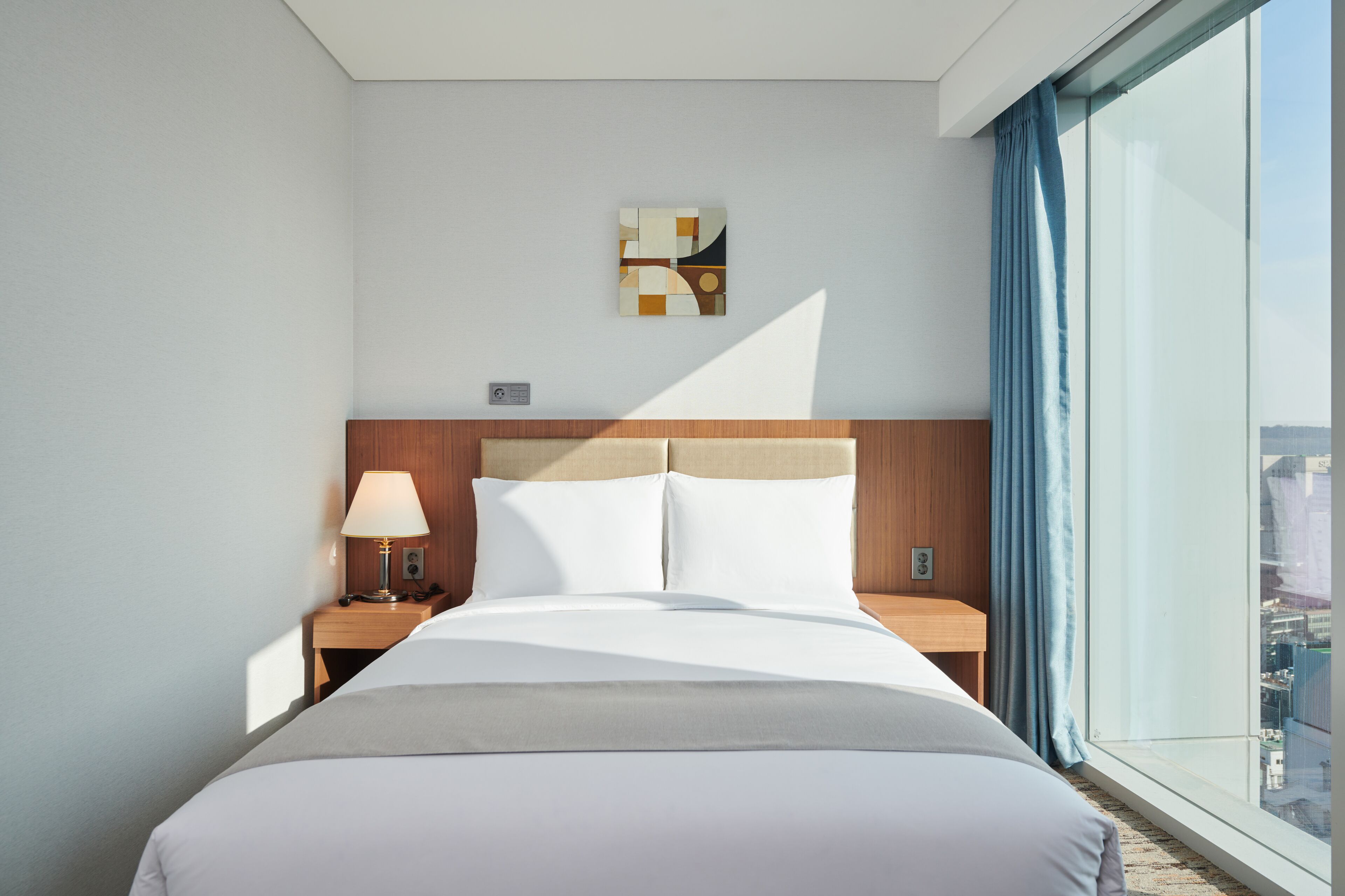 A hotel room with a large bed, a nightstand with a lamp, a wall-mounted artwork, and a view of the city through a large window.