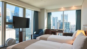 Habitación de hotel con dos camas, un escritorio, una silla, un televisor y ventanas amplias con vistas a la ciudad.
