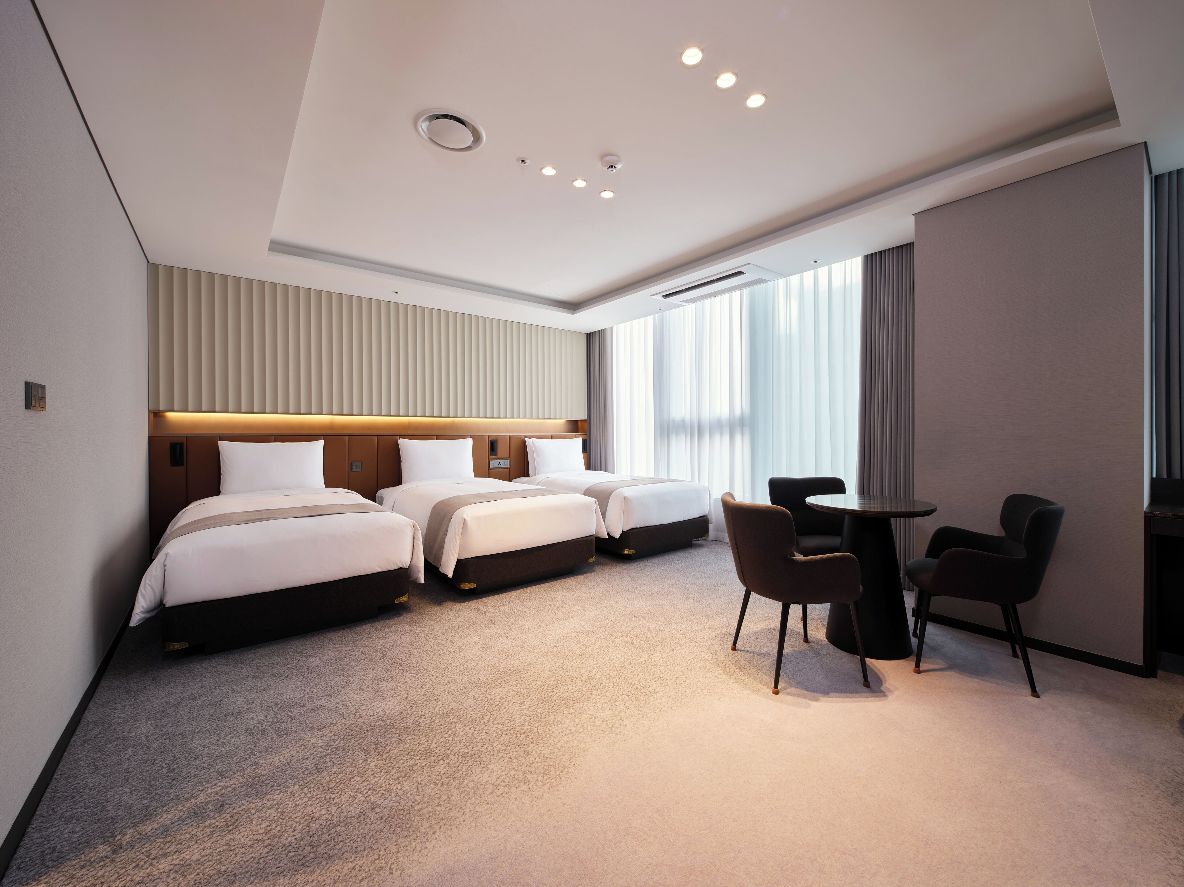 A hotel room with three beds, a small table, and chairs, a ceiling with recessed lighting, and a large window with curtains.