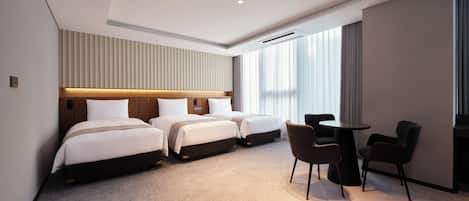 A hotel room with three beds, a small table, and chairs, a ceiling with recessed lighting, and a large window with curtains.