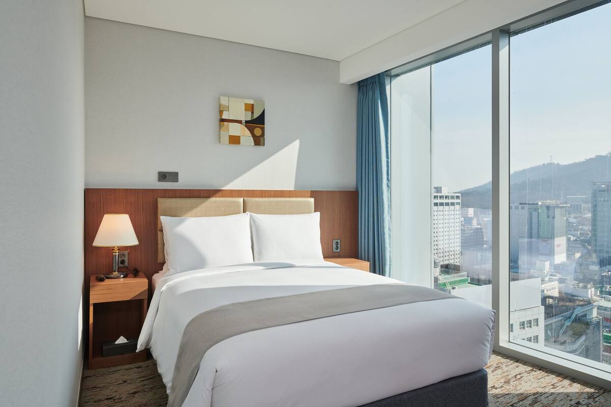 Ein Hotelzimmer mit einem großen Bett, einem Nachttisch mit Lampe, einem Bild an der Wand und Blick auf die Stadt durch das Fenster.