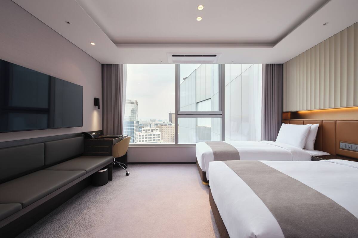 Ein modernes Hotelzimmer mit großem Fenster, einem Flachbildfernseher, einer Couch, zwei Betten und einem Schreibtisch.