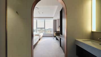 A modern hotel room with a large window, a bed, a desk, and a bathroom with a sink.
