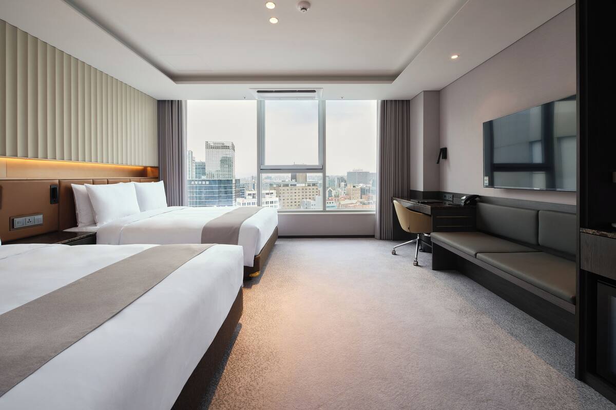 Ein Hotelzimmer mit zwei Betten, einem Schreibtisch, einem Fernseher und Blick auf die Stadt.