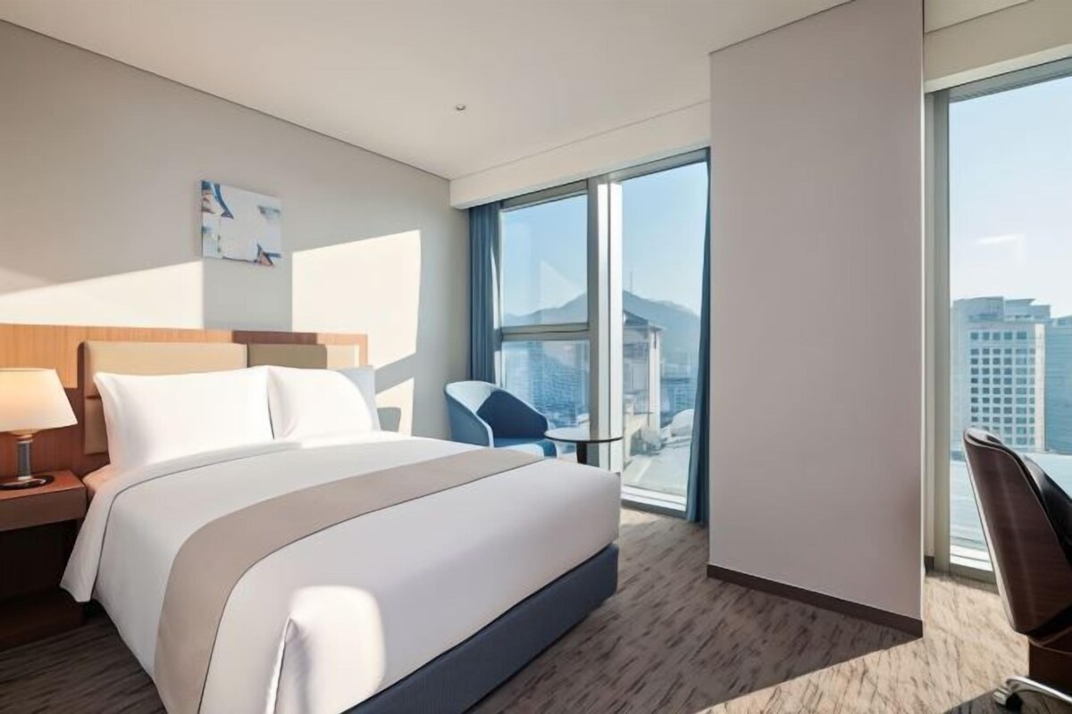 A modern hotel room with a large bed, a desk, a chair, and a view of the city through large windows.