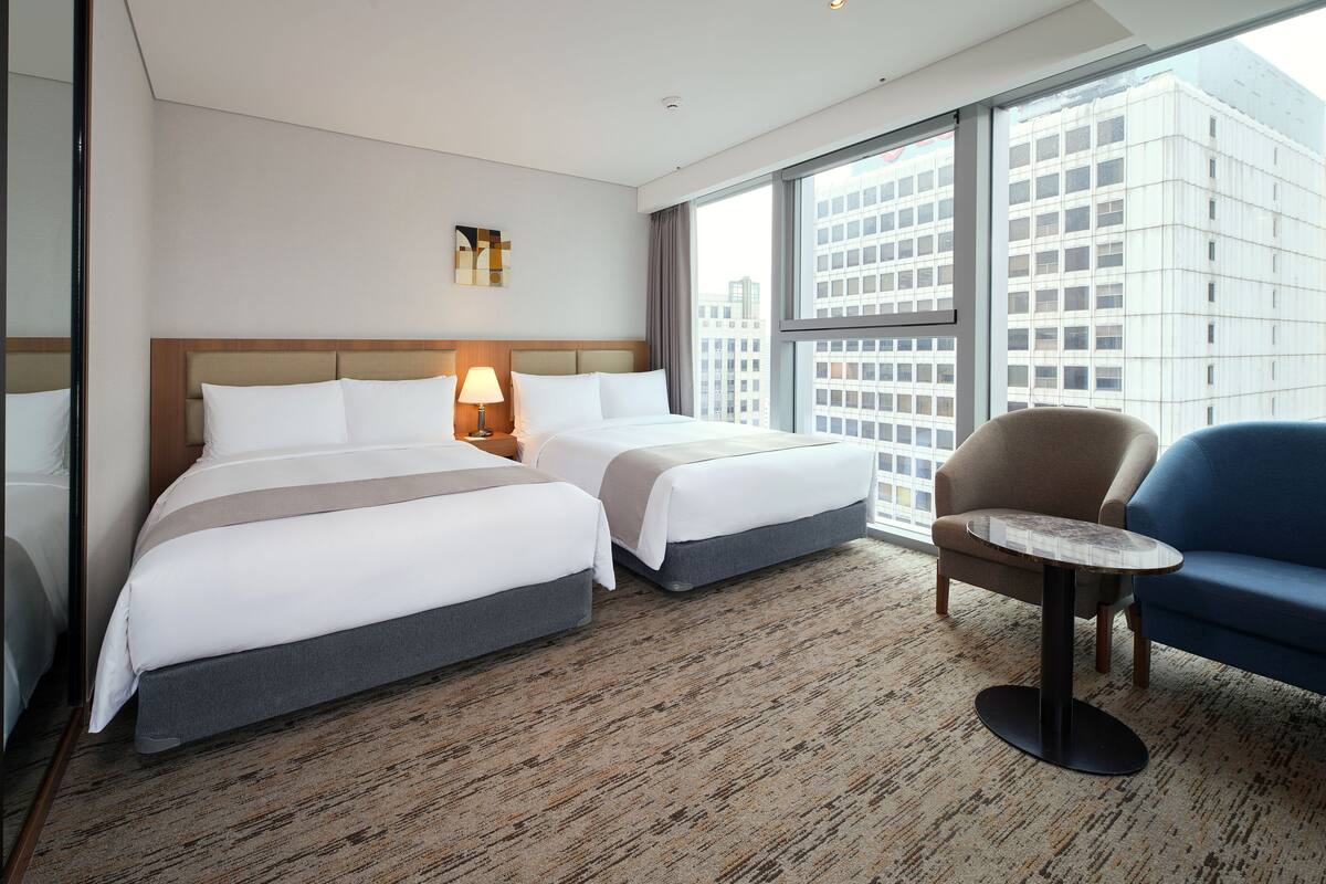 Ein Hotelzimmer mit zwei Betten, einem kleinen Tisch, einem Stuhl und großen Fenstern mit Blick auf die Stadt.