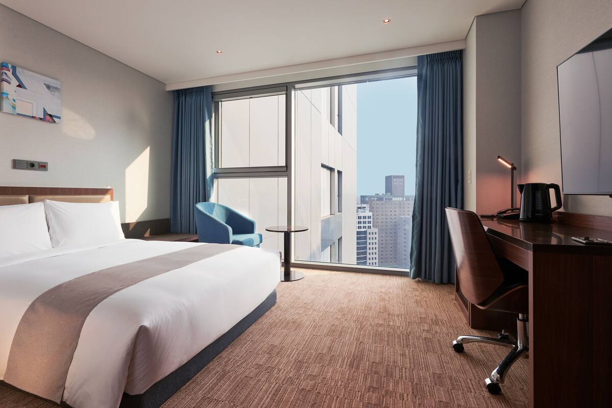 Ein Hotelzimmer mit einem großen Bett, einem Schreibtisch, einem Sessel, einem kleinen Tisch und einem großen Fenster mit Blick auf die Stadt.