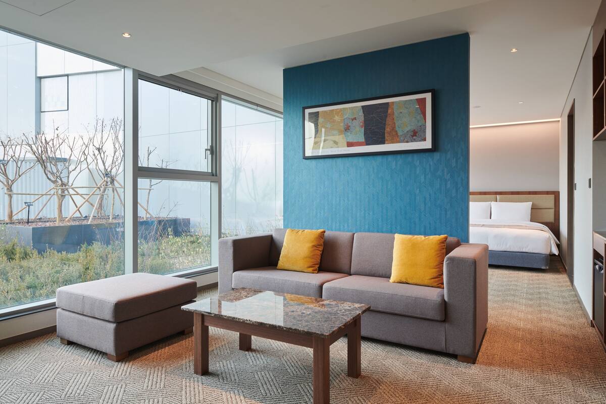 Ein modernes Hotelzimmer mit großem Fenster, einer gelb gepolsterten Couch, einem kleinen Hocker, einem Holztisch und einem Bett mit weißer Bettwäsche.