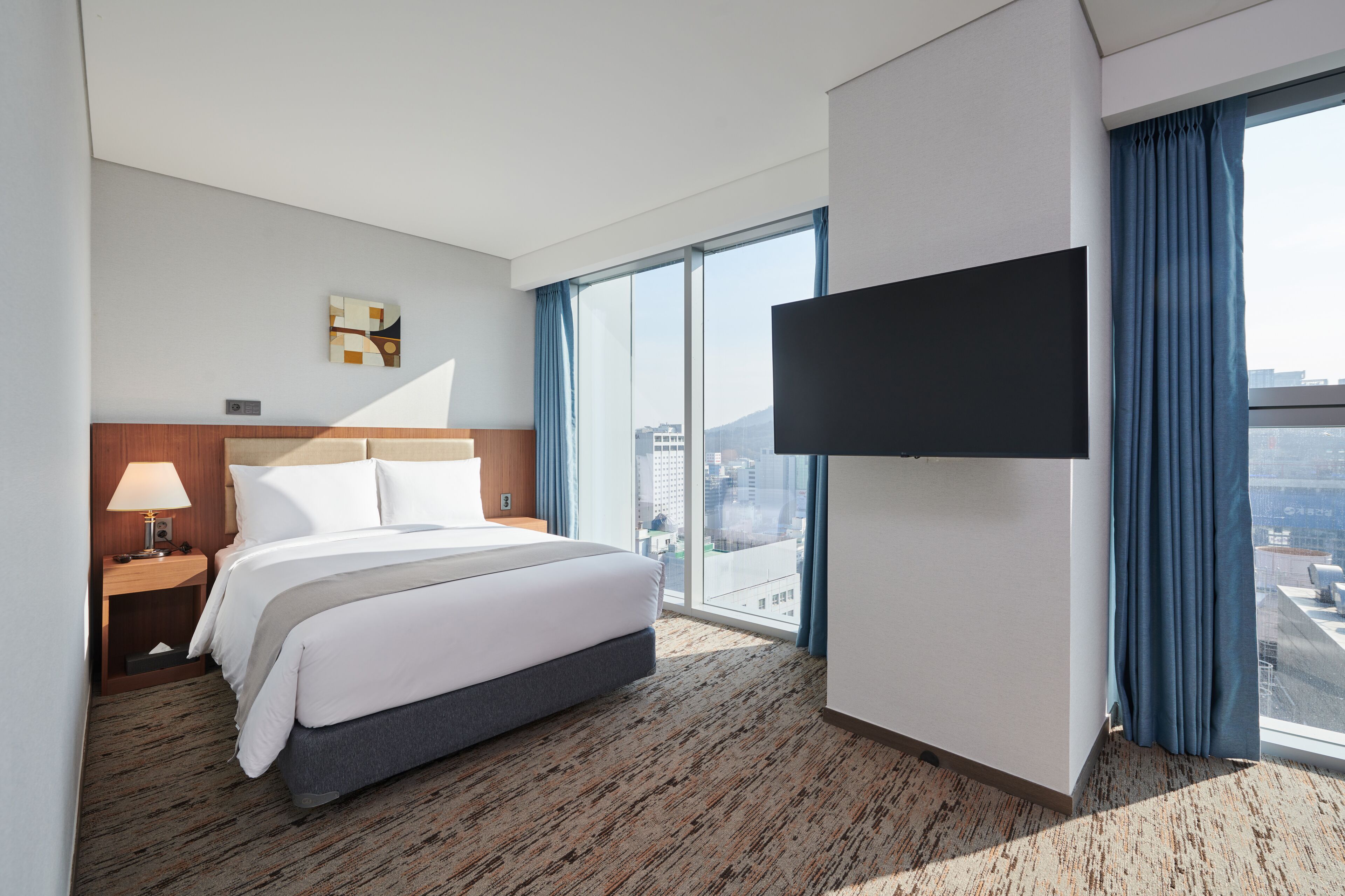 A hotel room with a large bed, a flat-screen TV mounted on the wall, and a view of the city through large windows.