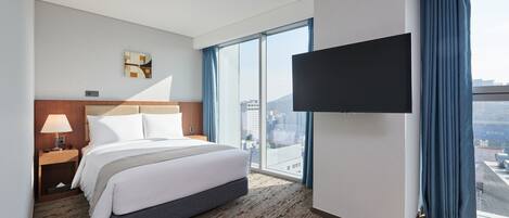 A hotel room with a large bed, a flat-screen TV mounted on the wall, and a view of the city through large windows.
