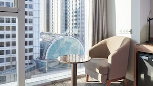 Una habitación de hotel moderna con un ventanal que ofrece vistas a un edificio de gran altura y una estructura de vidrio. Hay una mesa redonda y un sillón color beige.