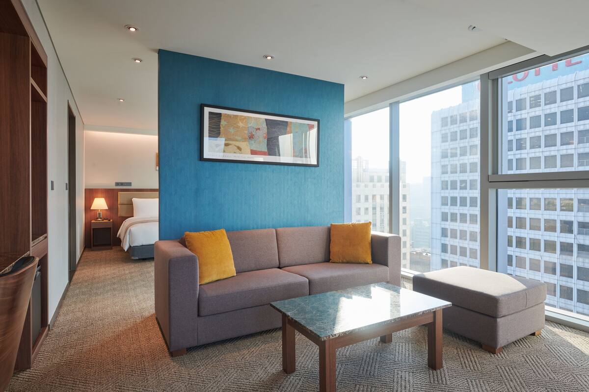 Ein modernes Hotelzimmer mit grauem Sofa, einem kleinen Tisch und einem großen Fenster mit Blick auf die Stadt.