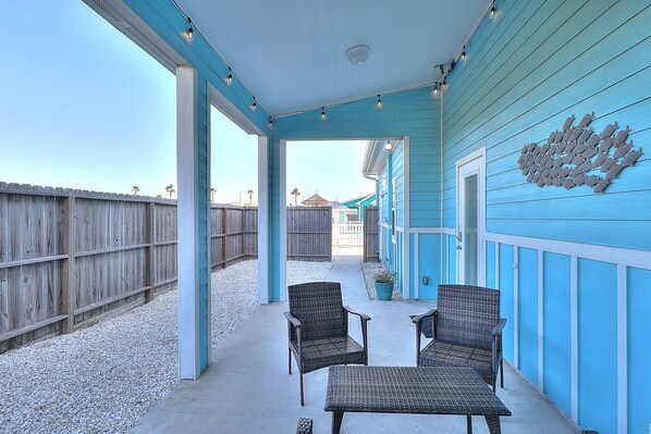 A covered patio with wicker furniture, a wooden fence, and a blue building with a decorative fish wall.