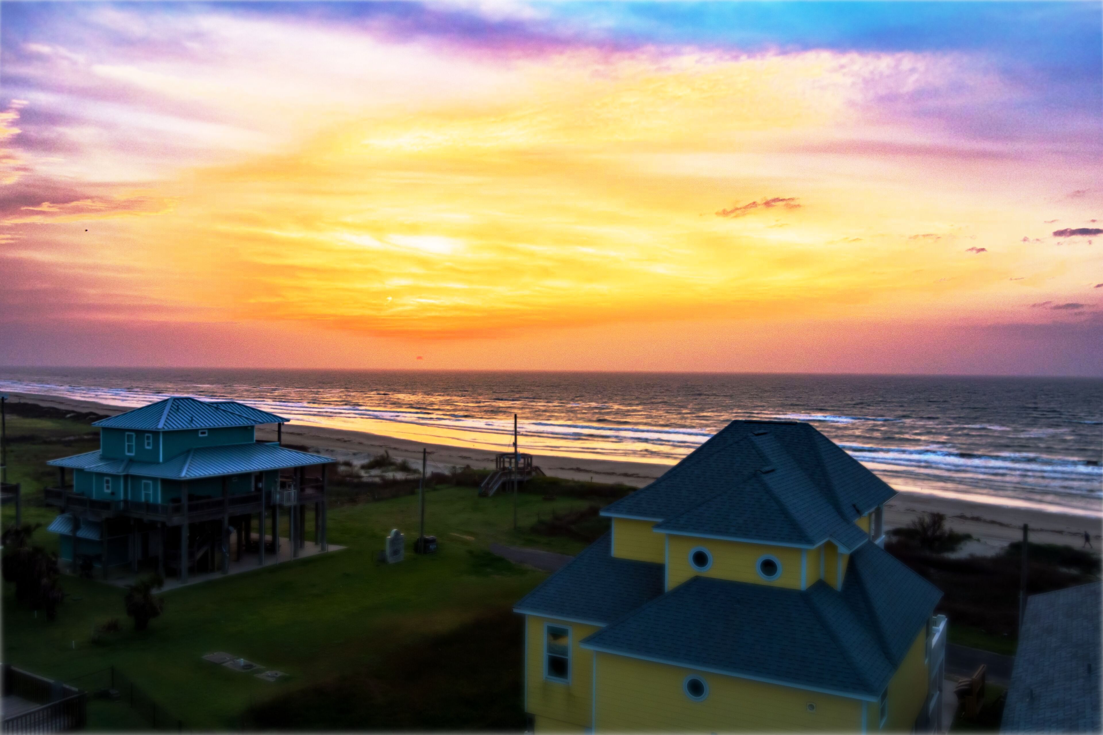 A coastal view with a sunset, featuring two houses with distinct architectural styles, one with a blue roof and the other with a yellow roof, overlooking the beach.