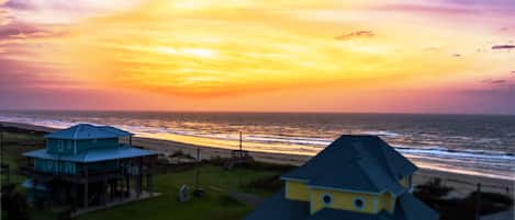 A coastal view with a sunset, featuring two houses with distinct architectural styles, one with a blue roof and the other with a yellow roof, overlooking the beach.