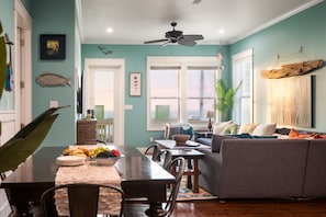 A dining area with a table set for a meal, a sofa, and a ceiling fan in a room with teal walls and wooden flooring. - New! Beachfront*Spectacular Views*4 bdrm House*Sleeps 14 (Crystal Beach)