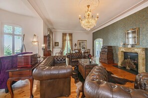 Ein geräumiges Wohnzimmer mit Ledersofas, einem prunkvollen Kronleuchter, einem Kamin und Blick ins Freie.