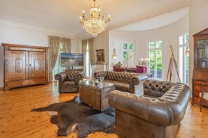 A spacious living room with leather sofas, a glass coffee table, a chandelier, and a large wooden cabinet.