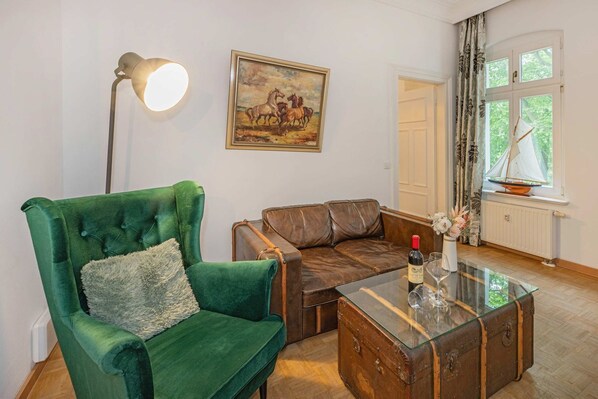 Una habitación con un sillón verde, un sofá de cuero marrón, una mesa de centro de vidrio con una botella y vasos, un cuadro de caballos y una ventana con cortinas.