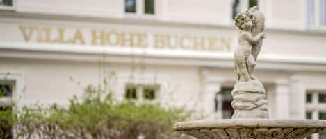 A stone fountain with a statue in front of a building with 'VILLA HOHE BUCHEN' signage.