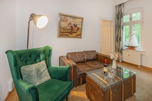 A room with a green armchair, a brown leather sofa, a glass coffee table with a bottle and glasses, a painting of horses, and a window with curtains. - Beautiful vacation apartment in Heringsdorf with garden, only 150 m to the sea - Villa (Heringsdorf)